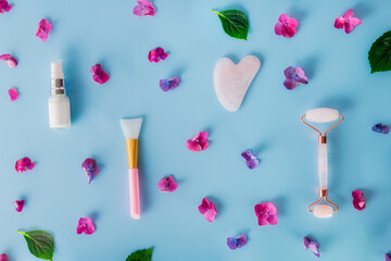 Top view composition with pink jade roller massager, gouache scraper, applying brush and skin serum on blue background with pink hydrangea flower petals. Anti-age, lifting, toning skin treatment.
