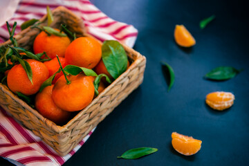 Fresh ripe mandarins, clementine, tangerine with green leaves in the straw bowl on a striped napkin and dark blue stone background. Local citrus fruit harvest. Selective focus.