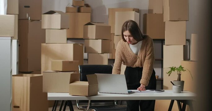 young woman is chatting by video call in laptop looking on screen and web camera in her home warehouse with boxes