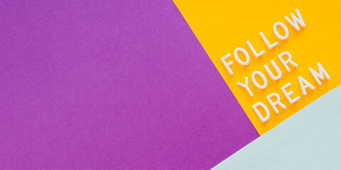 Diagonal banner with text FOLLOW YOUR DREAM on purple, yellow and light blue background. Top view on inspiring quote on colorful paper backdrop.