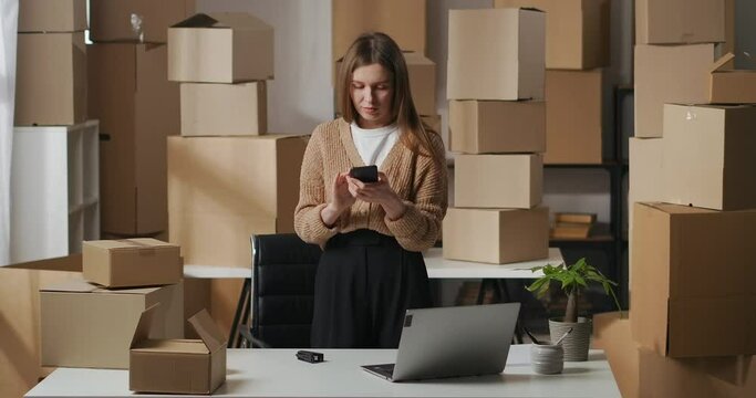 female business owner is checking email in smartphone in domestic warehouse, standing between cupboard boxes with goods