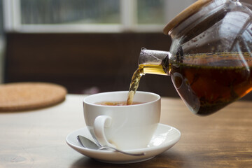 Tea from a glass teapot is poured into a white porcelain cup against the background of a wooden table. Shallow depth of field. Background blurred