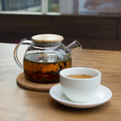 Glass teapot and white porcelain cup with tea on a wooden table. Shallow depth of field. Background blurred