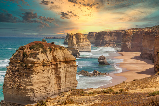 Blue green ocean and beach with sandbanks cliffs and waves  with close view of The twelve apostles and cliffs in the shadow of the sunset  in Victoria, Australia against a orange cloudy sky