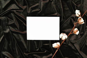 White invitation card mockup on a dark brown wavy velvet with a dried cotton branch. Flat lay
