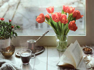 Oriental coffee in traditional Turkish copper coffee pot with flowers on window sill. Wooden windowsill with bunch of tulips book. Cold rainy day in spring. Cozy scene, hygge concept.