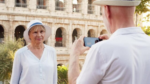 Happy tourists family in hats enjoy vacation in Rome. Senior man take photo of his wife on mobile phone near famous Italian attraction Colosseum. Seniors having fun travelling together