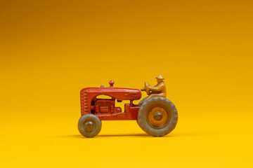 Small red vintage agrarian agricultural tractor with figurine on top. Studio toy still life against a seamless yellow background.