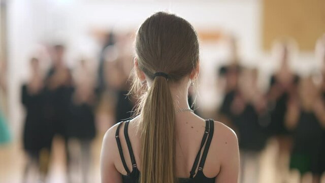 Back view close-up of Caucasian girl standing in dancing school with blurred children clapping at background. Confident talented brunette child receiving ovations in dance studio. Art and success.