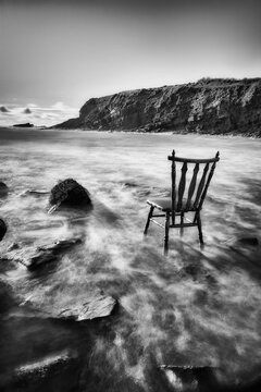 vintage antique chair tucked into the water of an Irish beach surrounded by rocks and cliffs. long exposure with traces of water. mono image