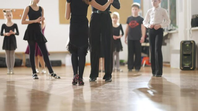 Unrecognizable couple of teens rehearsing in dance studio with children watching around. Confident fit boy and girl walking in slow motion to start position in classical dance.