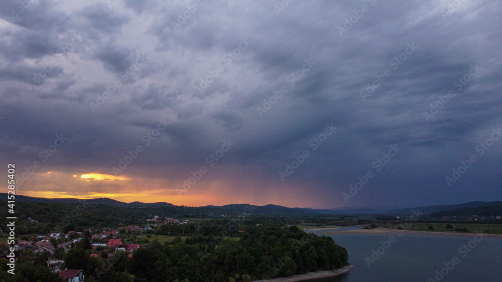 Poster Aerial shot of a lake with buildings and mountains under the cloudy sky at sunset