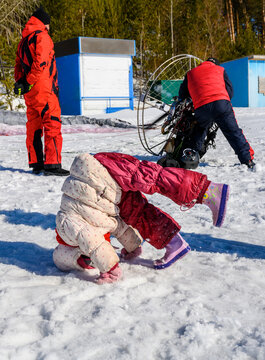 A child happily tumbles in the snow against the background of adults engaged in setting up a motor paraplane