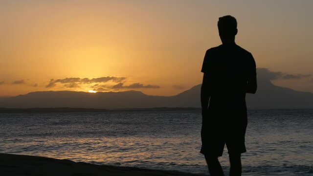 Homme debout devant un superbe couch&eacute; de soleil en face de la mer. Le soleil se couche derri&egrave;re des montagnes.