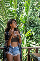 Portrait of young brunette woman freelancer drinking coffee from white cup on balcony of tropical bungalow with palm trees view