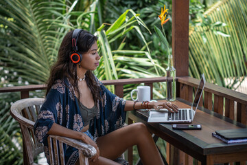 Portrait of young brunette woman freelancer, hands in bracelets, in necklace with shell working on laptop, smartphone, headphones on table, on balcony of tropical bungalow with palm trees view