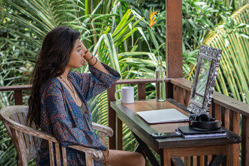 Portrait of young brunette woman looking to the mirror, hands in bracelets, necklace with shell on balcony of tropical bungalow with palm trees view