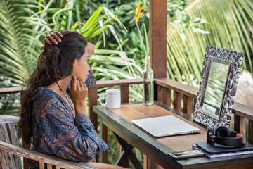 Portrait of young brunette woman looking to the mirror, hands in bracelets holding earring on balcony of tropical bungalow with palm trees view