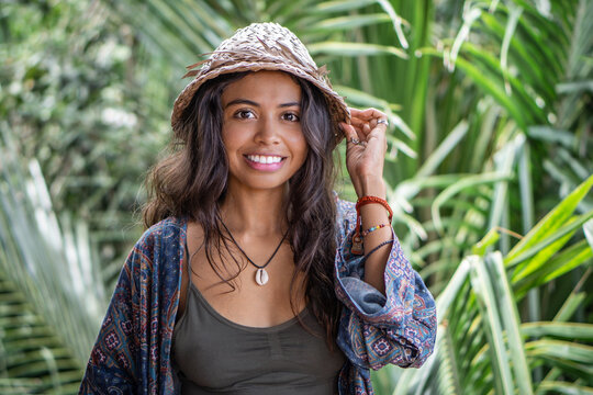 Portrait of young brunette woman in a straw hat, hands in bracelets, in necklace with shell on balcony of tropical bungalow with palm trees view