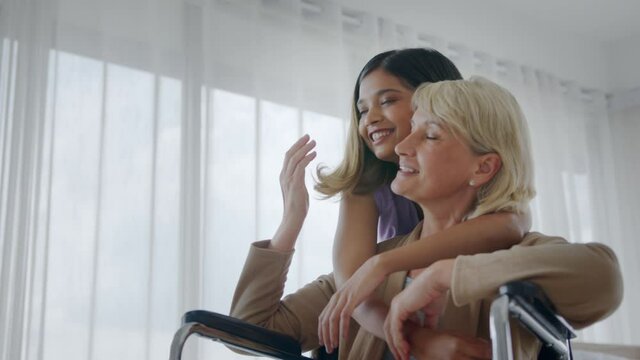 Medium close up - Joyful adopted Asian teen daughter comforting and embracing her injured Caucasian stepmother sitting in wheelchair. Love and caring time of biracial family concept.
