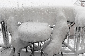 Snow-covered summer furniture on the balcony through ice drops on the glass on winter cloudy day