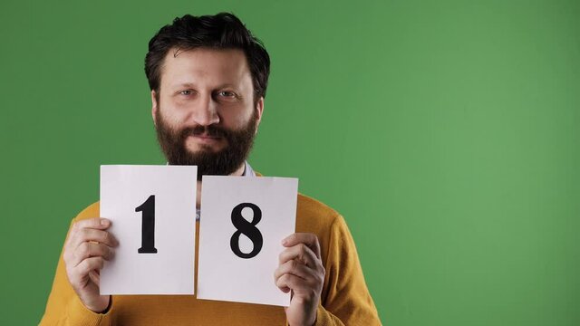 Man holding number 18 sign. Cheerful white bearded good-looking man in yellow sweater shows number eighteen on white sheet. Chrome key, green screen, copy space