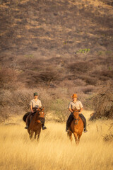 Two women ride through grass by hill