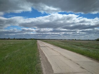 straight road ahead among the green grass in the clouds in the sky in summer travel