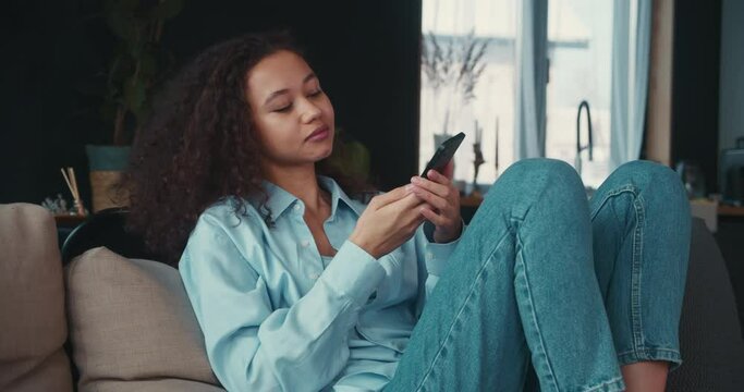 Smart phone addiction. Happy young smiling mixed race student woman with curly hair using shopping app at home on sofa.