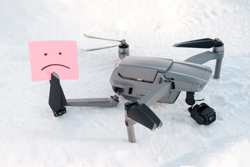 Sticky note with sorry smile in propeller blades. Broken gimbal camera and drone motor arm after crash on snow in winter outdoor closeup. Expensive repairs concept
