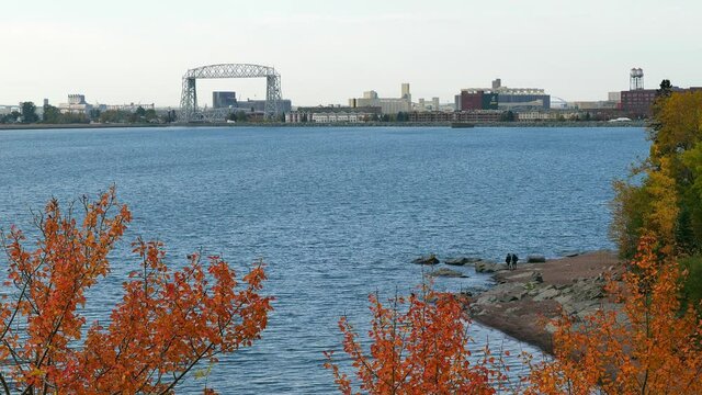 View of iconic Duluth Minnesota Aerial Lift Bridge, water and shore of Lake Superior, nearby autumn leaves and distant harbor buildings on a cloudy fall day.