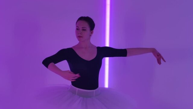 A young slender ballerina in a white tutu performs graceful pas with arms and legs. Studio rehearsal against the backdrop of multi-colored neon lights. Slow motion. Close up.