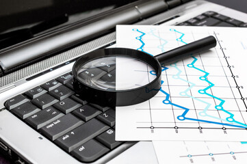 Magnifying glass with financial graphs on the laptop keyboard. Business concept.