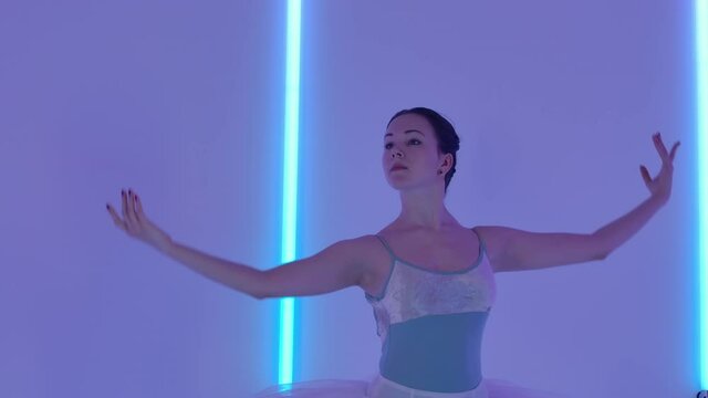 Professional ballerina dancing ballet In the studio against the background of multicolored neon tubes. A girl in a stage outfit works out graceful pas with her hands and jumps. Slow motion. Close up.