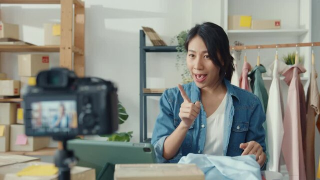 Young Asia lady fashion designer using mobile phone receiving purchase order and showing clothes recording video live streaming online at shop. Small business owner, online market delivery concept.