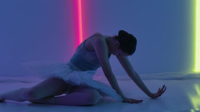 Ballerina demonstrates her flexibility in the studio against a backdrop of colorful neon lights. The dancer, sitting on the floor, works out pas with her hands and does a split. Slow motion. Close up.