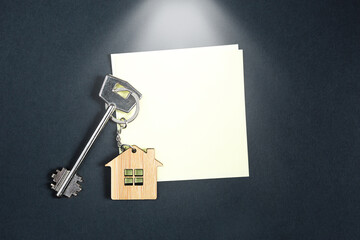 Keychain in the shape of wooden house with key on a black background with a square sheet for notes. Building, design, project, moving to new house, mortgage, rent and purchase real estate. Copy space