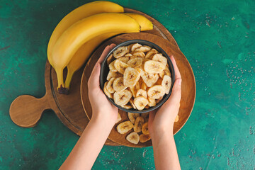 Woman holding bowl with crispy banana chips on color background