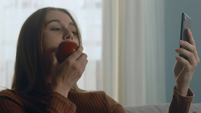 Young woman in blue jeans and brown sweater sits on beige sofa on window background, holding red apple and taking selfie. Close-up of a static plan.
