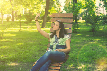 Cheerful girl after shopping with many shopping bags sitting on bench in the park and taking selfie with smart phone. Young woman takes a selfie, sits on a bench in a city park. toned
