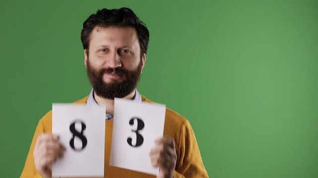 Man holding number 83 sign. Cheerful white bearded good-looking man in yellow sweater shows number eighty-three on white sheet. Chrome key, green screen, copy space