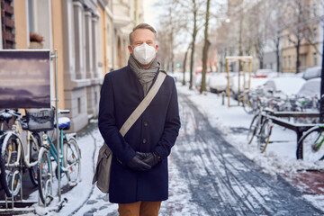Businessman wearing a protective surgical face mask during the Covid-19 or coronavirus pandemic and winter overcoat with leather bag over his shoulder standing in a snowy urban street 