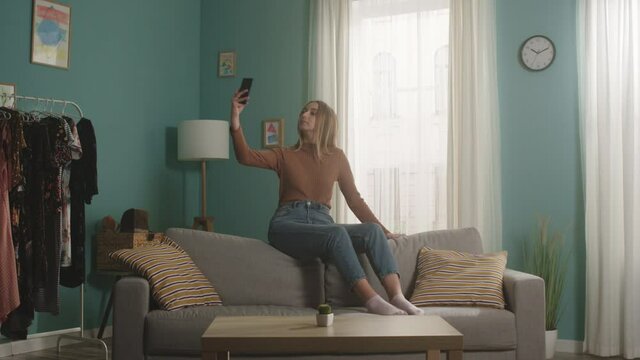 Woman in blue jeans and brown sweater sitting on the back of the sofa on the background of the window, taking a selfie. There is a cactus on the coffee table. General static plan.