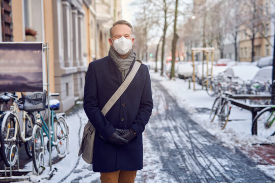Businessman wearing a protective surgical face mask during the Covid-19 or coronavirus pandemic and winter overcoat with leather bag over his shoulder standing in a snowy urban street - Powered by Adobe