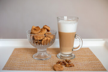 Homemade cookies and walnuts are in a bowl on a white table next to a glass glass of cappuccino. Close-up