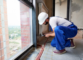 Fototapeta premium Handsome plumber connecting radiator to house heating system with a wrench, next to the window. Side view. Copy space. Installation process. Concept of plumbing works and home renovation.