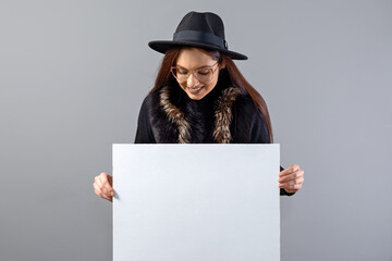 elegant young woman in glasses and hat showing empty board banner, isolated on gray, copy space