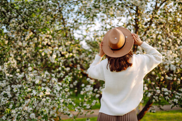 Portrait of Beautiful  Woman In hat  posing near flowering tree. Smiling young woman enjoying smell of flowers on background of spring garden.  Fashion, lifestyle. 