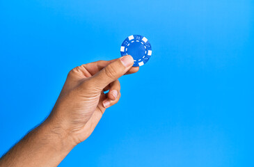 Hand of hispanic man holding casino chip over isolated blue background.