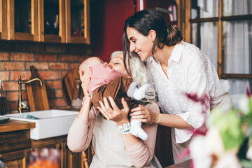 Joyful aged lady plays with little granddaughter sitting in daughter hugs near brown wooden kitchen.